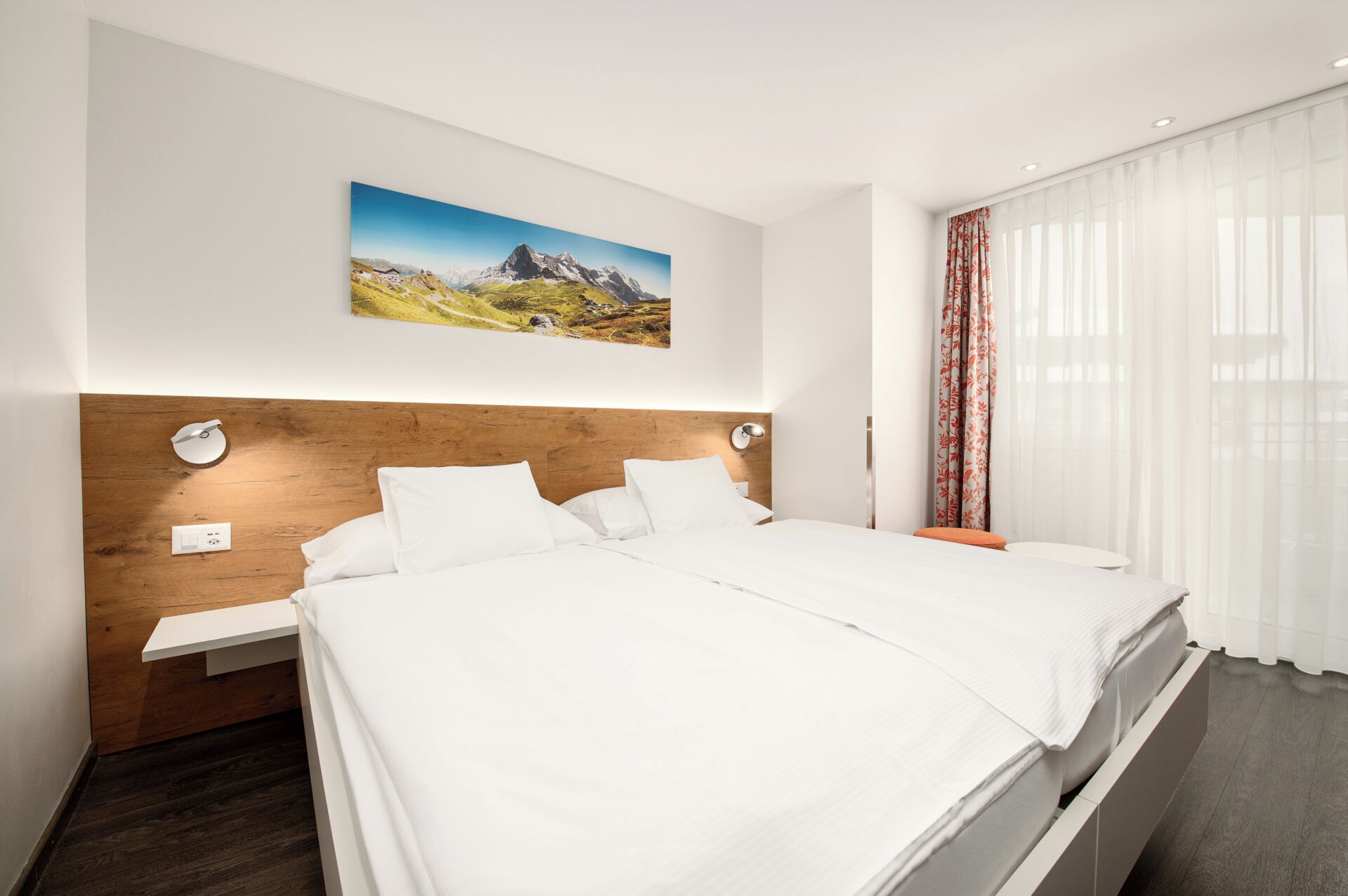 Una habitación de hotel moderna con cabecera de madera, una cama grande con sábanas blancas, una mesita de noche, una lámpara de pared, un cuadro de un paisaje montañoso y cortinas translúcidas.