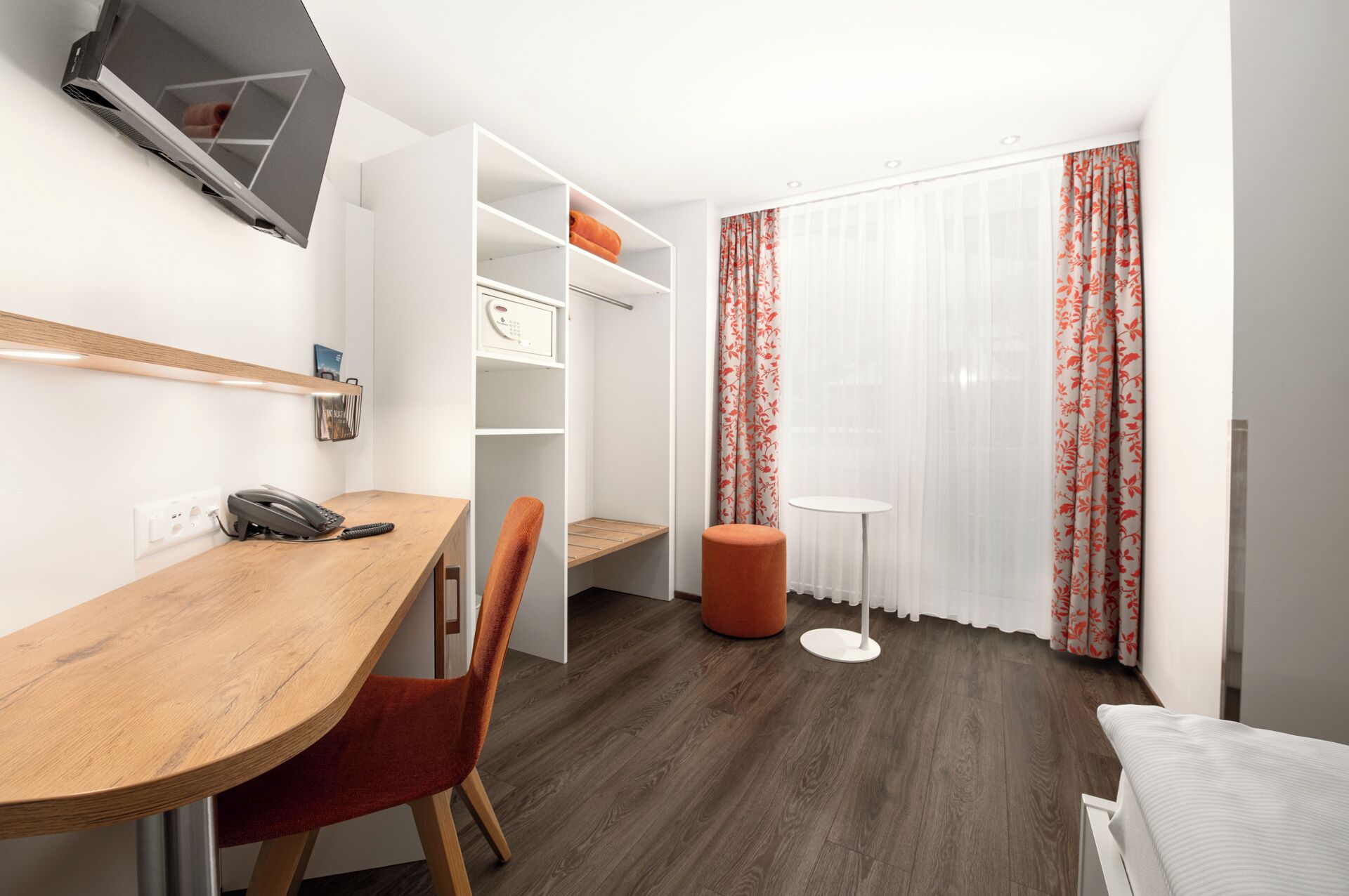 A hotel room with a wooden desk, a chair, a small round ottoman, a white round table, a wardrobe, and a window with floral curtains.