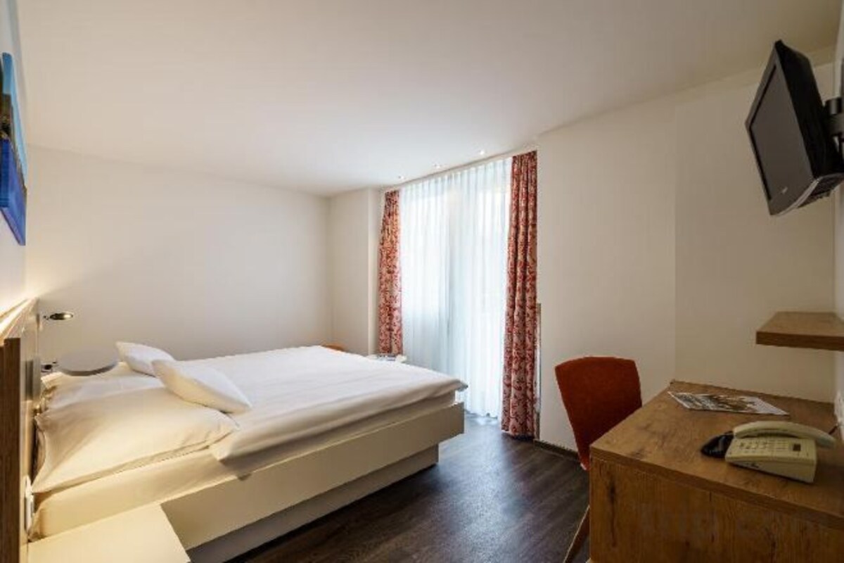 A hotel room with a bed, a desk, a chair, a TV, and a window with curtains.