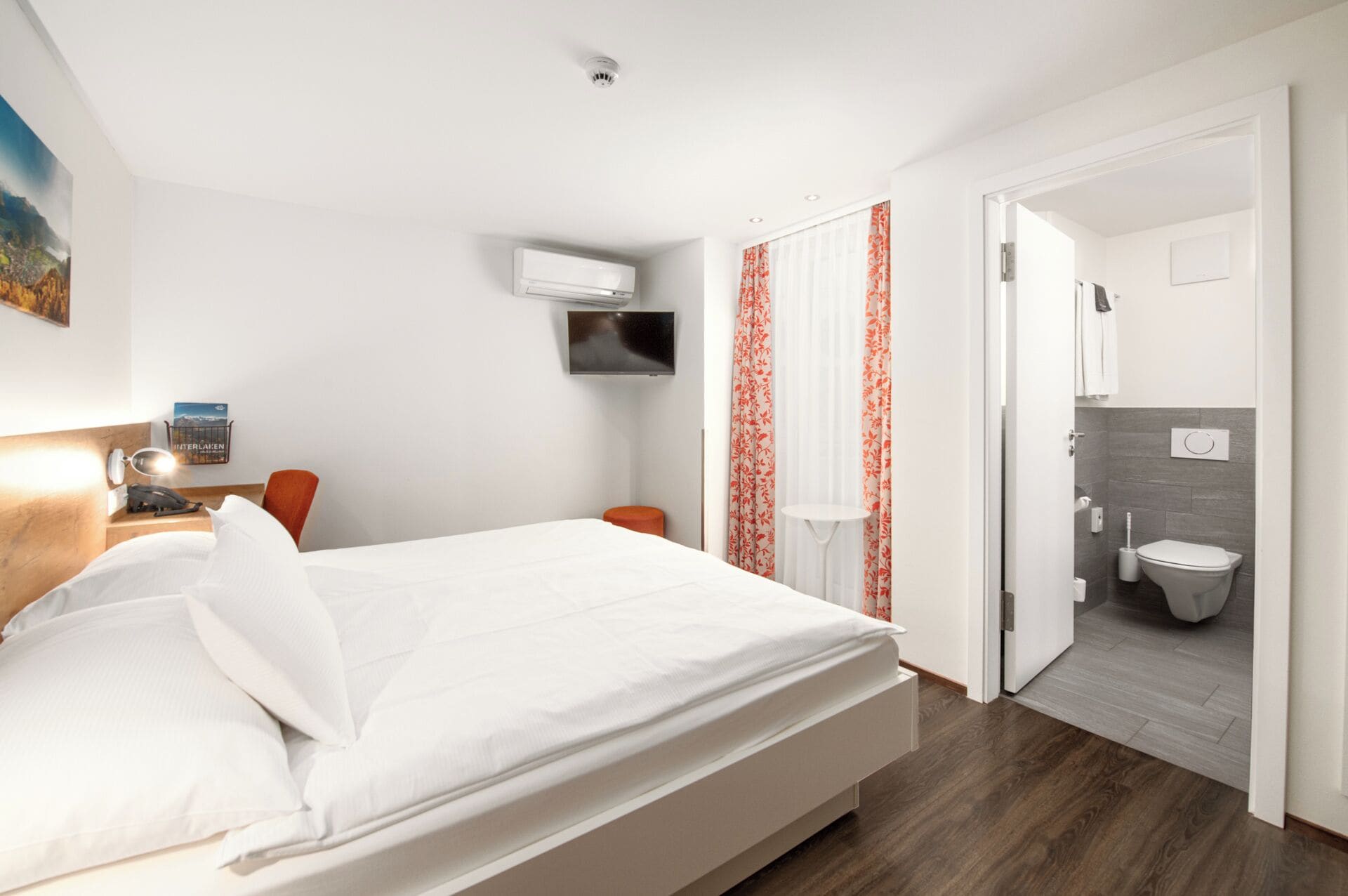 A hotel room with a bed, a TV mounted on the wall, a small table with a lamp, and a view into the bathroom.