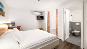 A hotel room with a bed, a TV mounted on the wall, a small table with a lamp, and a view into the bathroom.