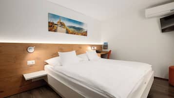 Une chambre d’hôtel moderne avec une tête de lit en bois, un lit avec du linge de maison blanc, un tableau accroché au mur, une table de chevet avec une lampe et un climatiseur fixé au mur.