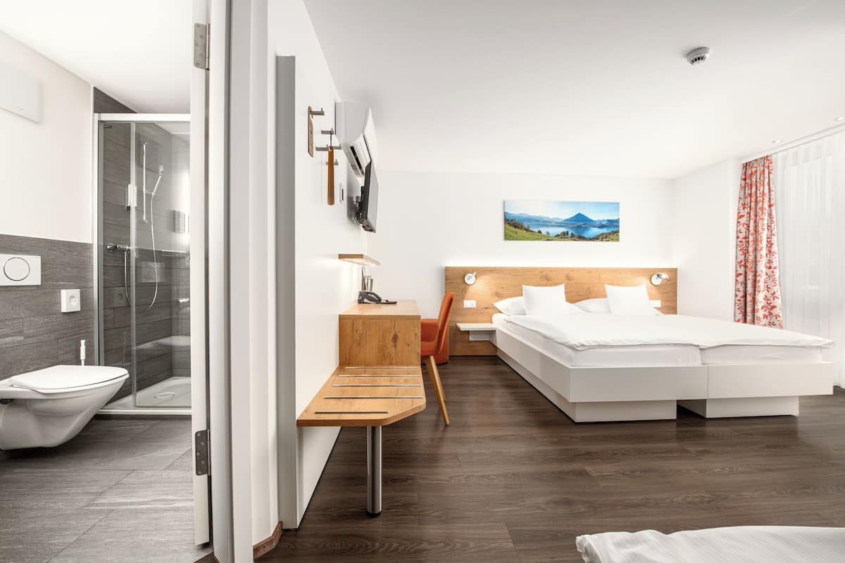A modern hotel room with a shower, toilet, wooden desk, bed, and a painting on the wall.