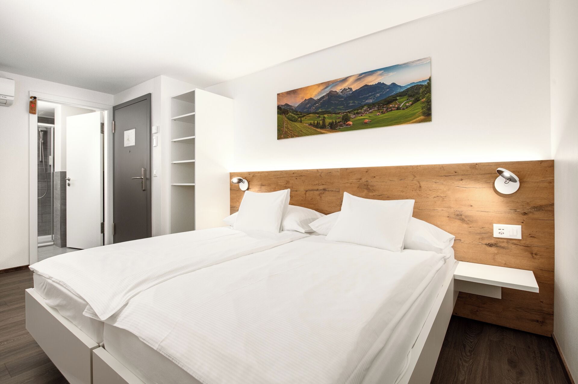 Una habitación de hotel moderna con cabecera de madera, cama con sábanas blancas, un cuadro colgado de un paisaje montañoso y una pequeña mesita de noche con una lámpara.