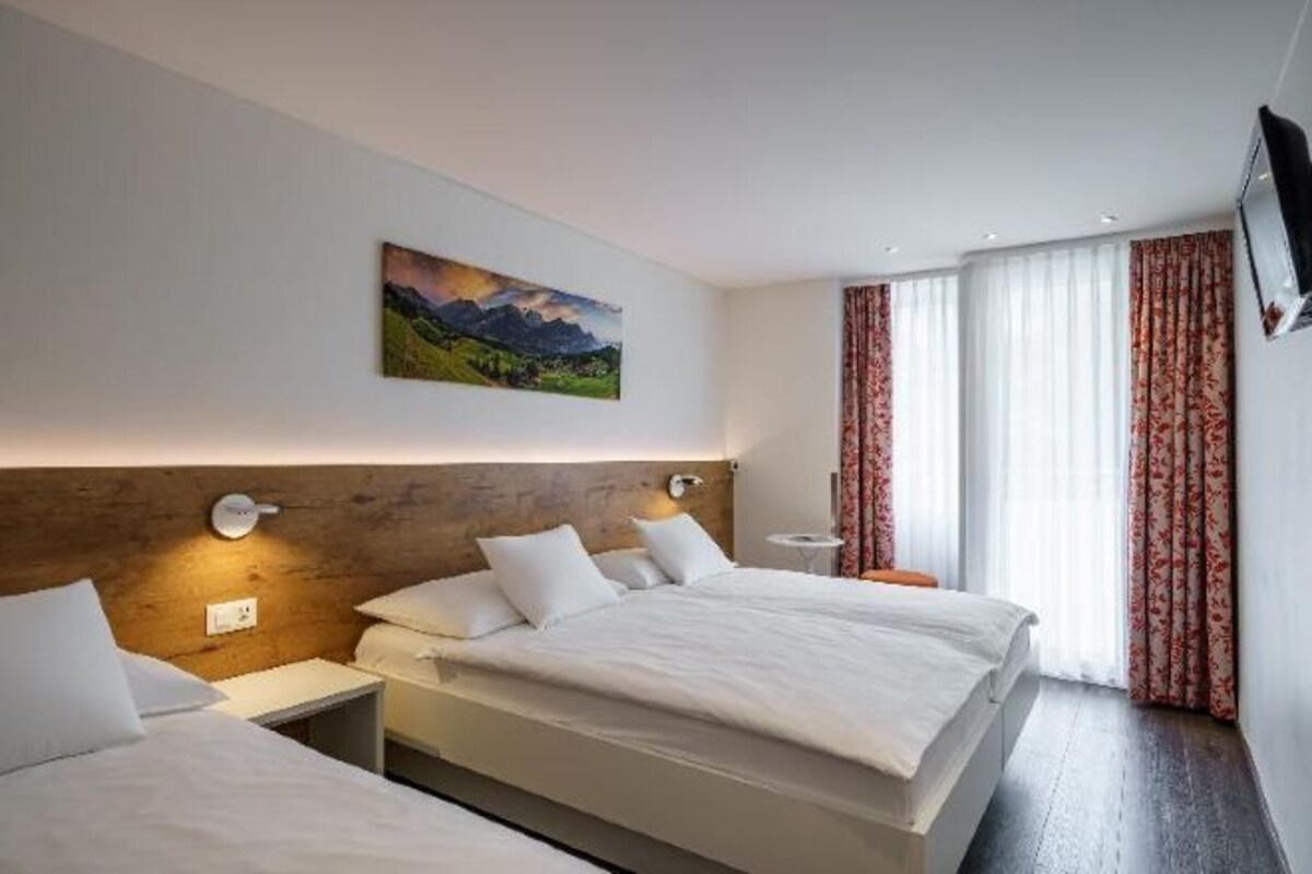 A hotel room with two beds, a painting on the wall, a TV mounted on the wall, and a window with curtains.