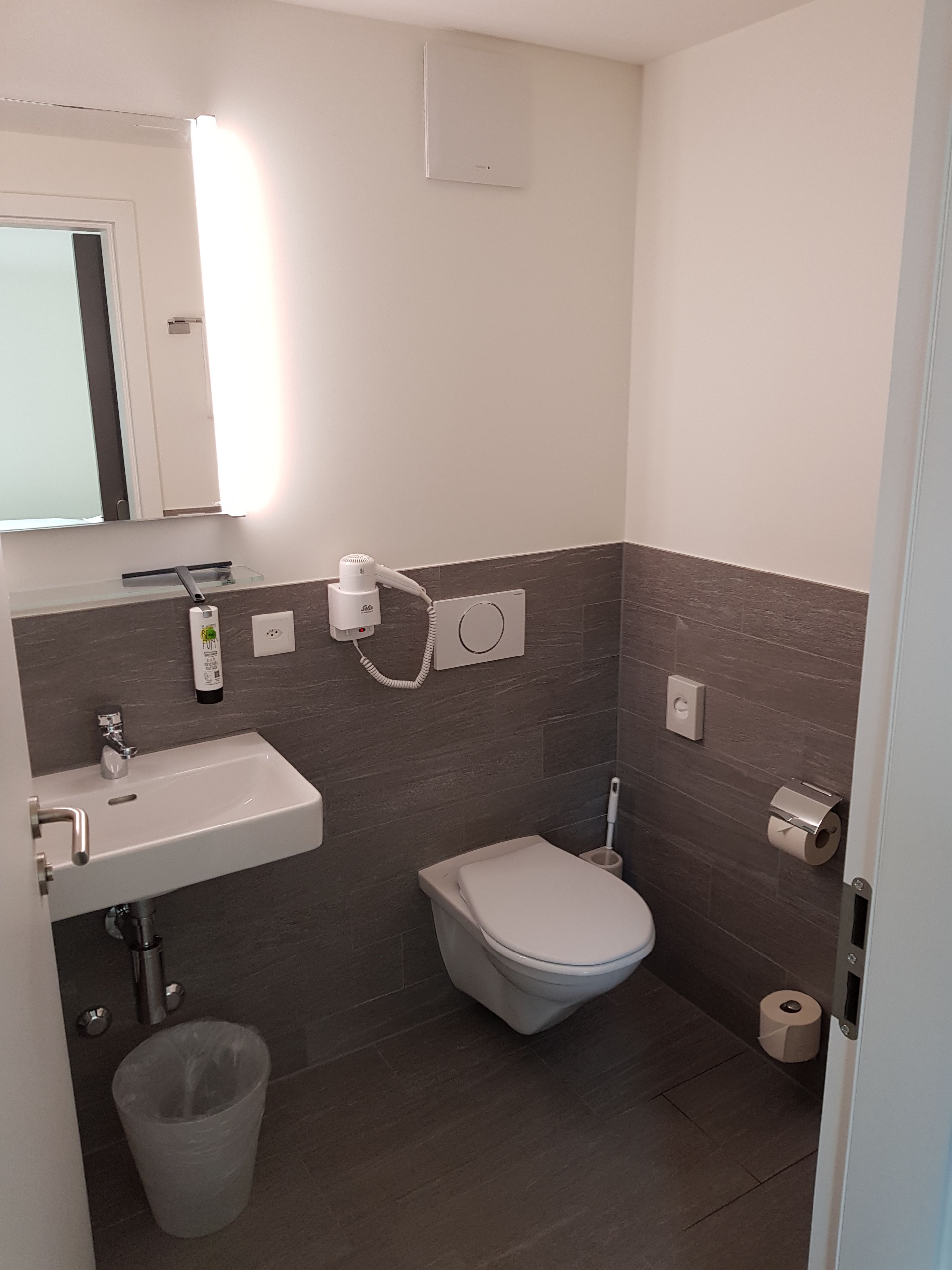 Un baño pequeño con un lavamanos blanco, un inodoro, un cubo de basura y un secador de pelo montado en la pared.