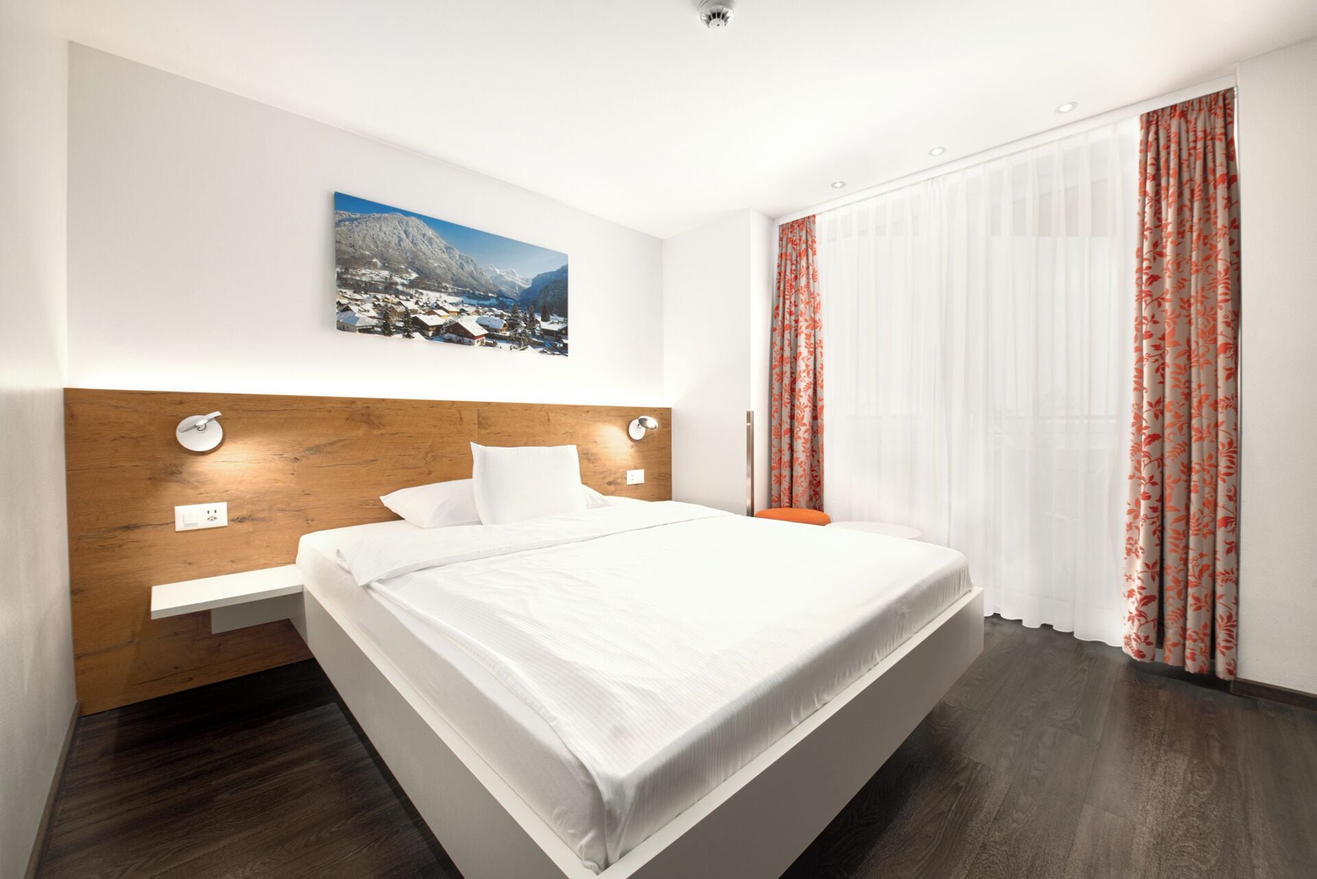 A modern hotel room with a wooden headboard, a large bed, a nightstand, a wall-mounted picture, and a window with sheer curtains.