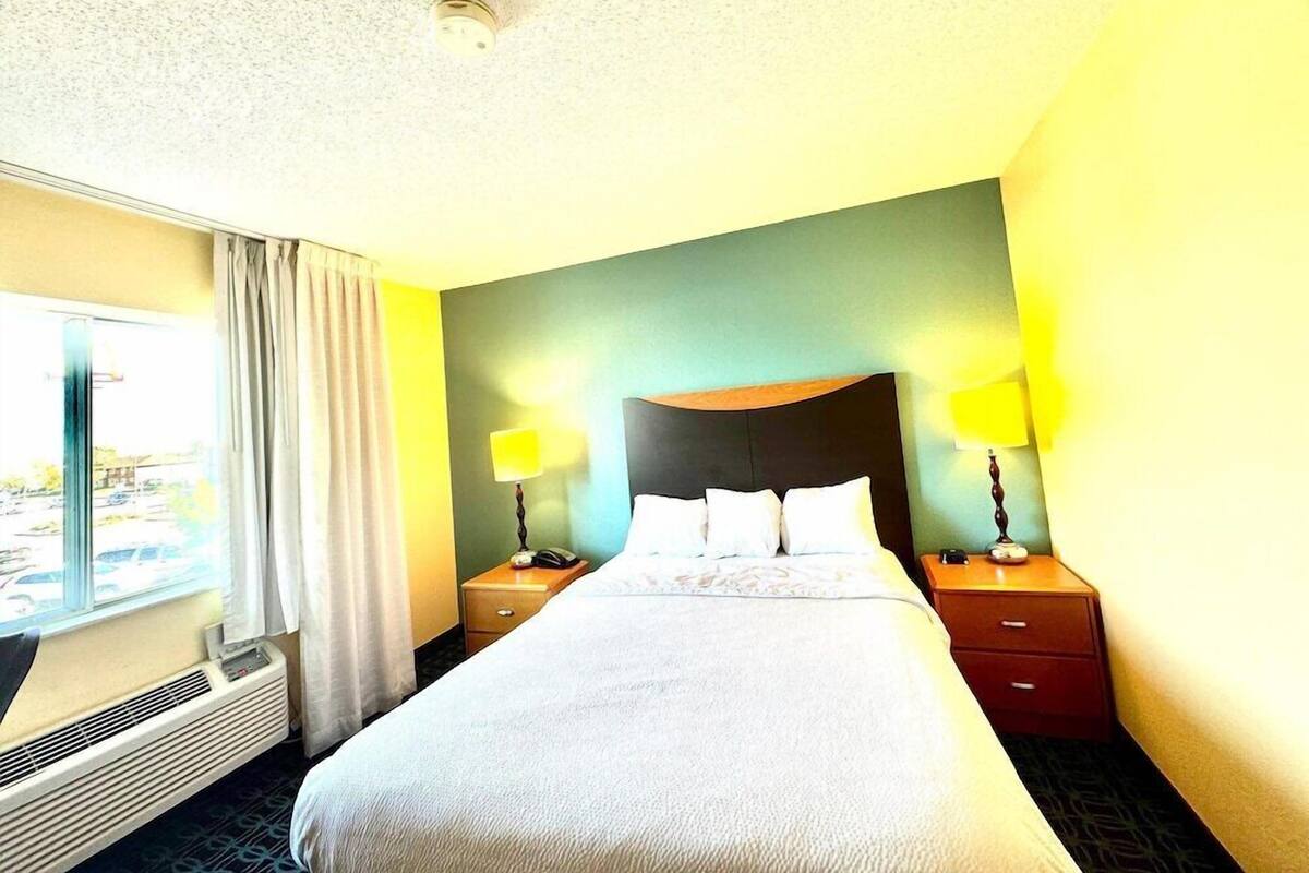A hotel room with a bed, two bedside tables, a window with curtains, and a wall-mounted heater.