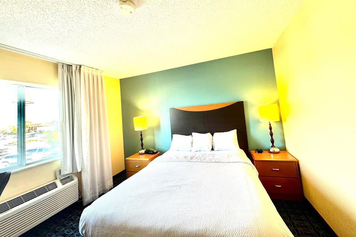 A hotel room with a large bed, two bedside tables with lamps, a window with curtains, and a ceiling vent.