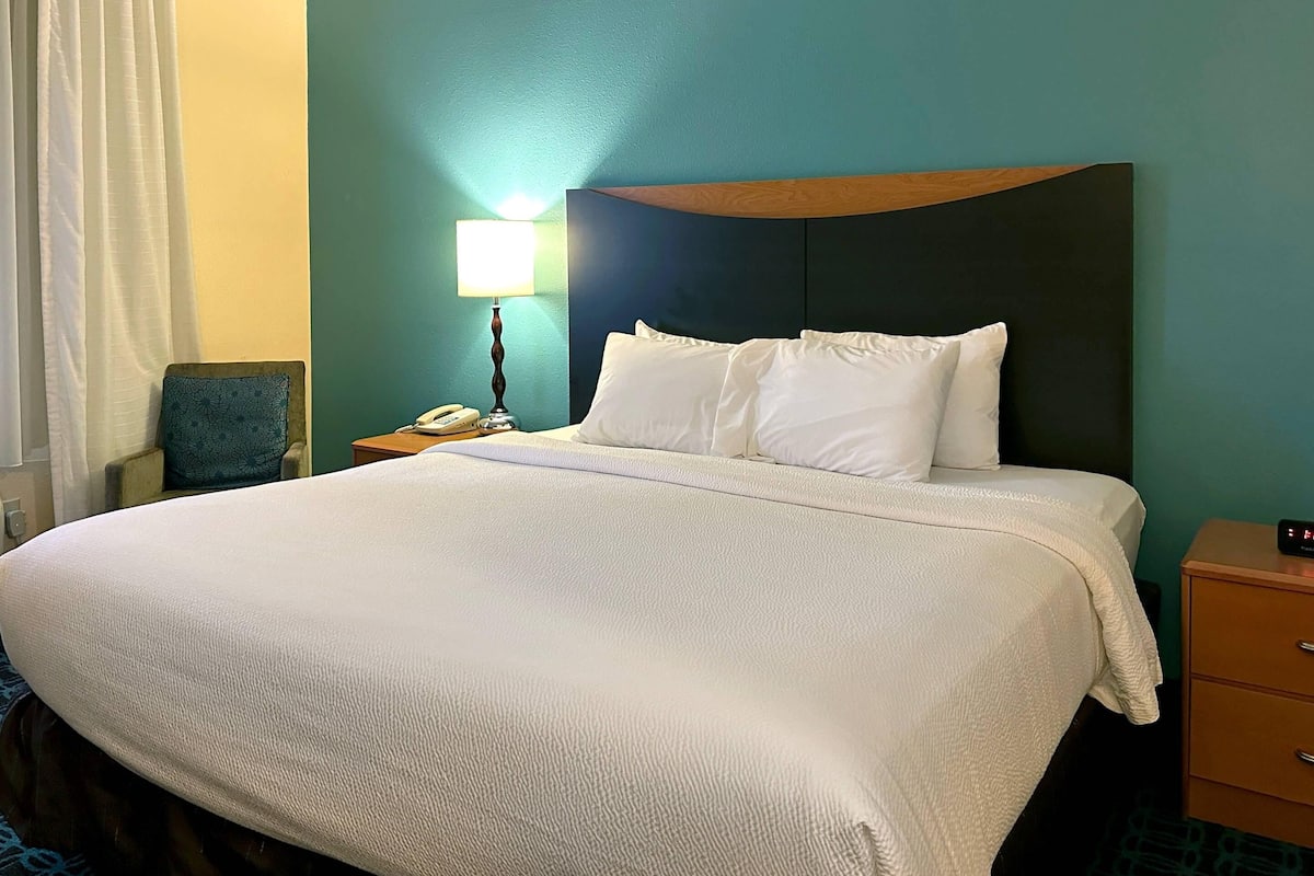 A hotel room with a large bed, a bedside table with a lamp, a chair, and a nightstand with a phone.