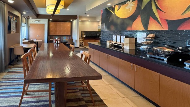 A modern hotel dining area with a long wooden table, cushioned chairs, and a kitchen counter with a fruit mural. - Country Inn & Suites by Radisson, Bismarck-Mandan (Bismarck)