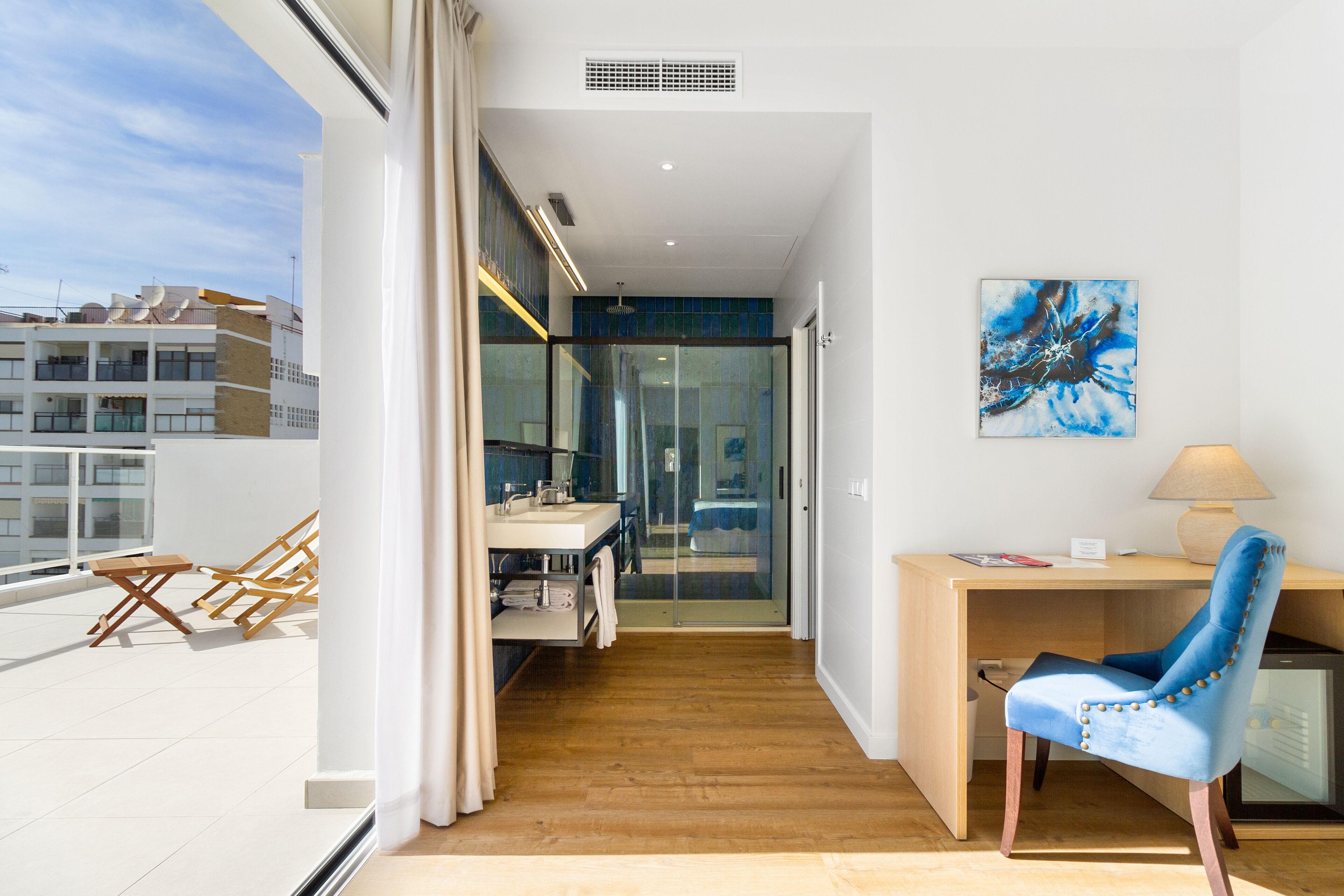 Una habitación moderna con un escritorio de madera, una silla azul, ducha de vidrio y vistas a la ciudad.