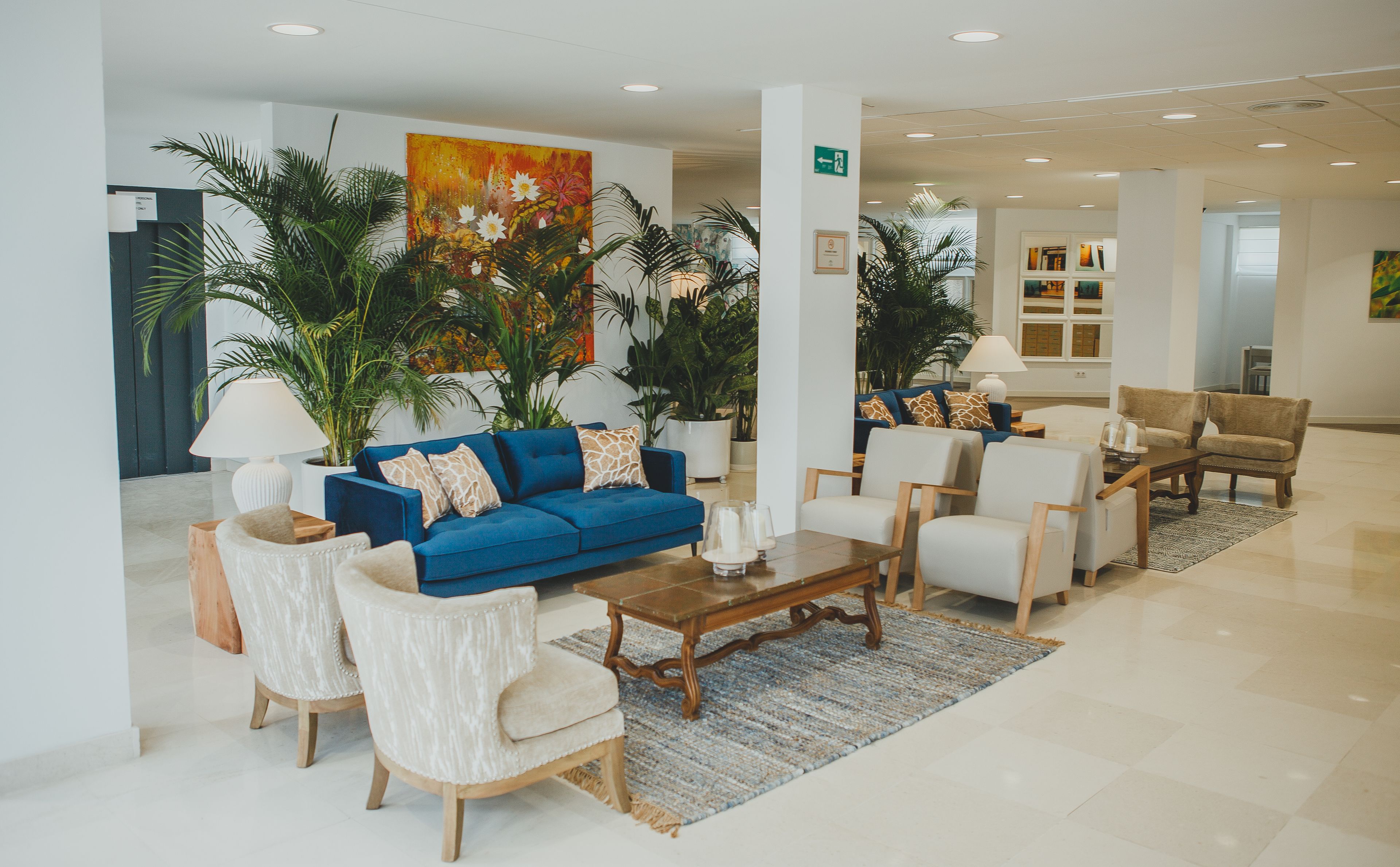 A spacious lobby with a blue sofa, white armchairs, and a coffee table. There are potted plants and a large painting on the wall.