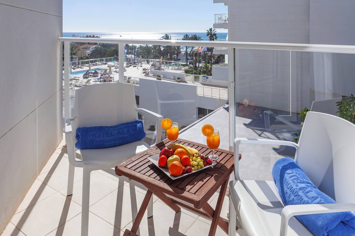 A balcony with a table set with fruits and juice, white chairs, and a view of the beach and pool.