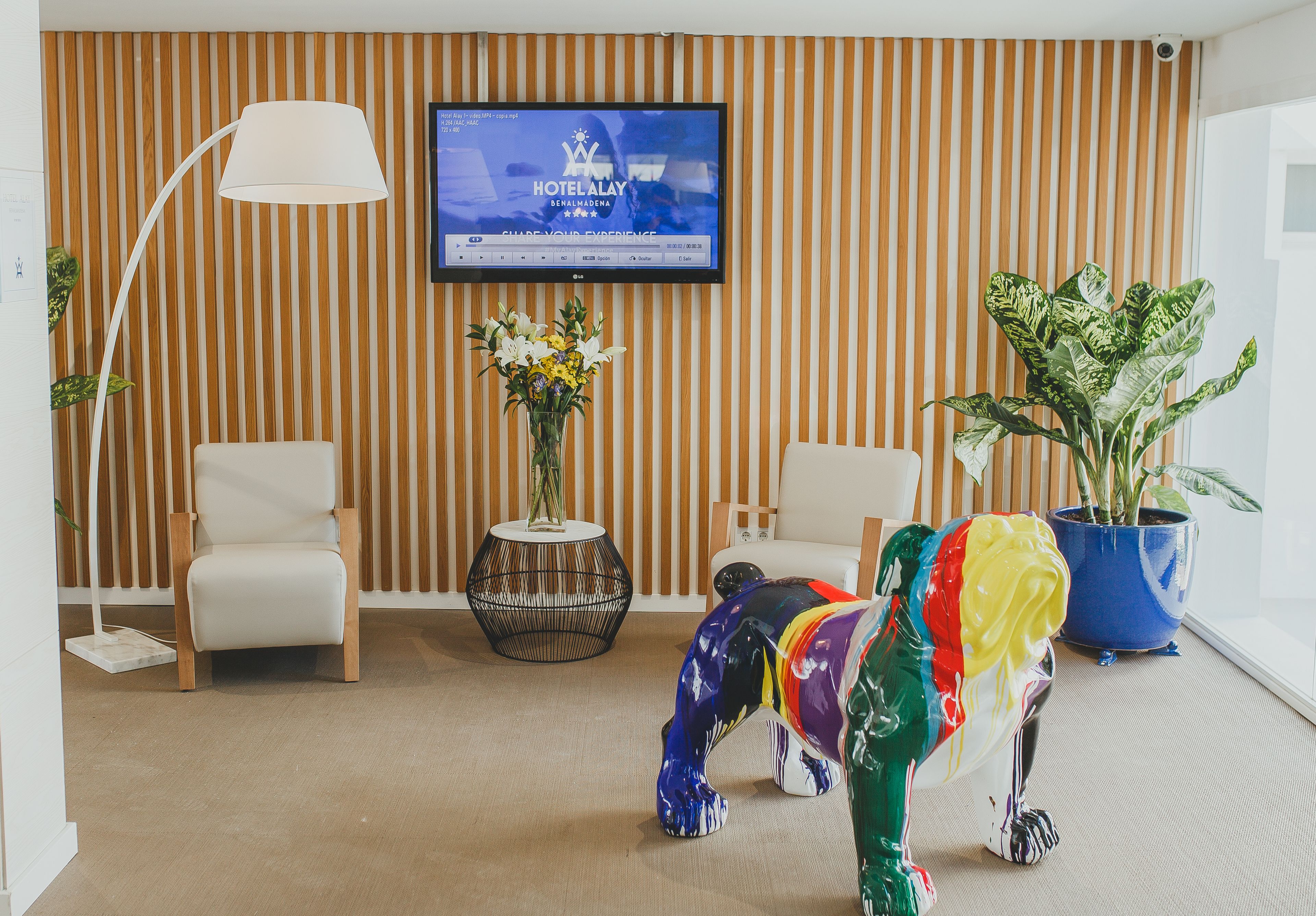 En moderne hotellobby med stribede trævægge, et fladskærms-tv der viser hotellets navn, to hvide lænestole, et rundt sofabord med en vase blomster og en farverig elefant skulptur.