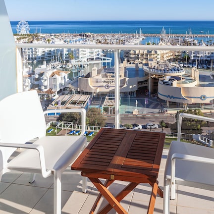 A balcony with two white chairs and a wooden table, overlooking a marina with boats and buildings.