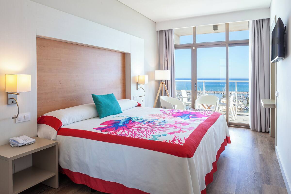 A hotel room with a large bed, a desk, a chair, a TV, and a balcony with a view of the sea.