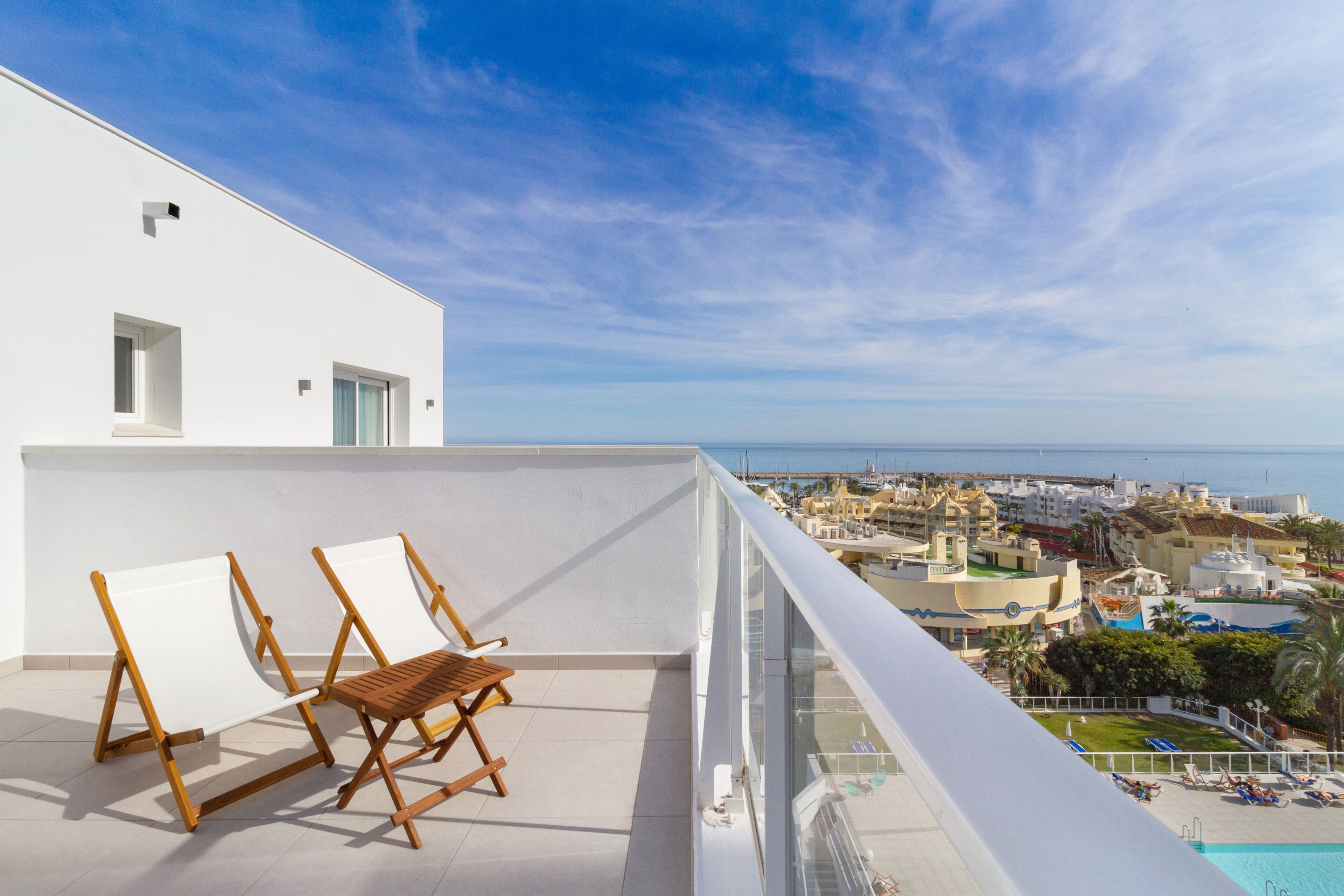 Een balkon met twee houten loungestoelen, een glazen balustrade en uitzicht op zee en de stad.