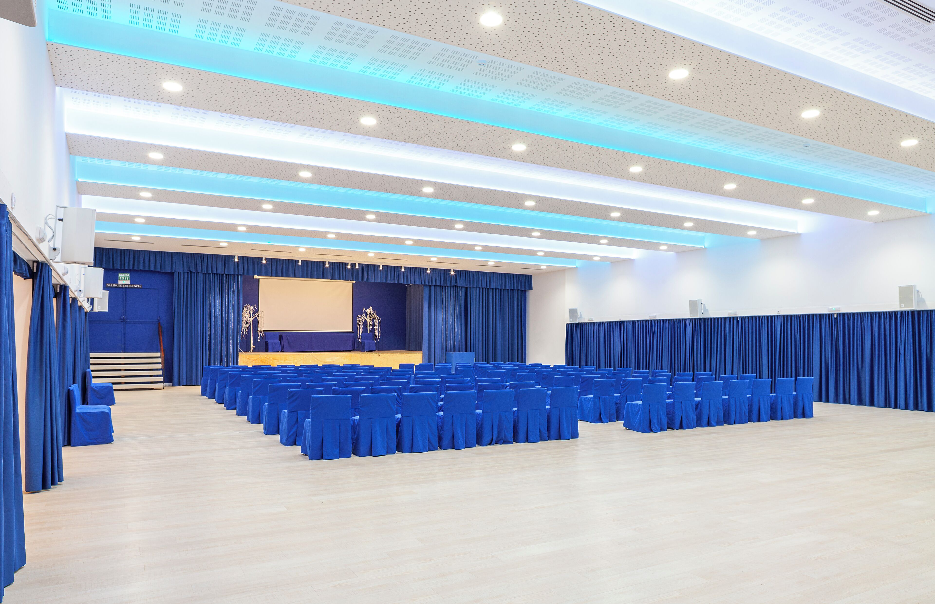 A conference room with rows of blue chairs, a stage with a blue backdrop, and a ceiling with recessed lighting.