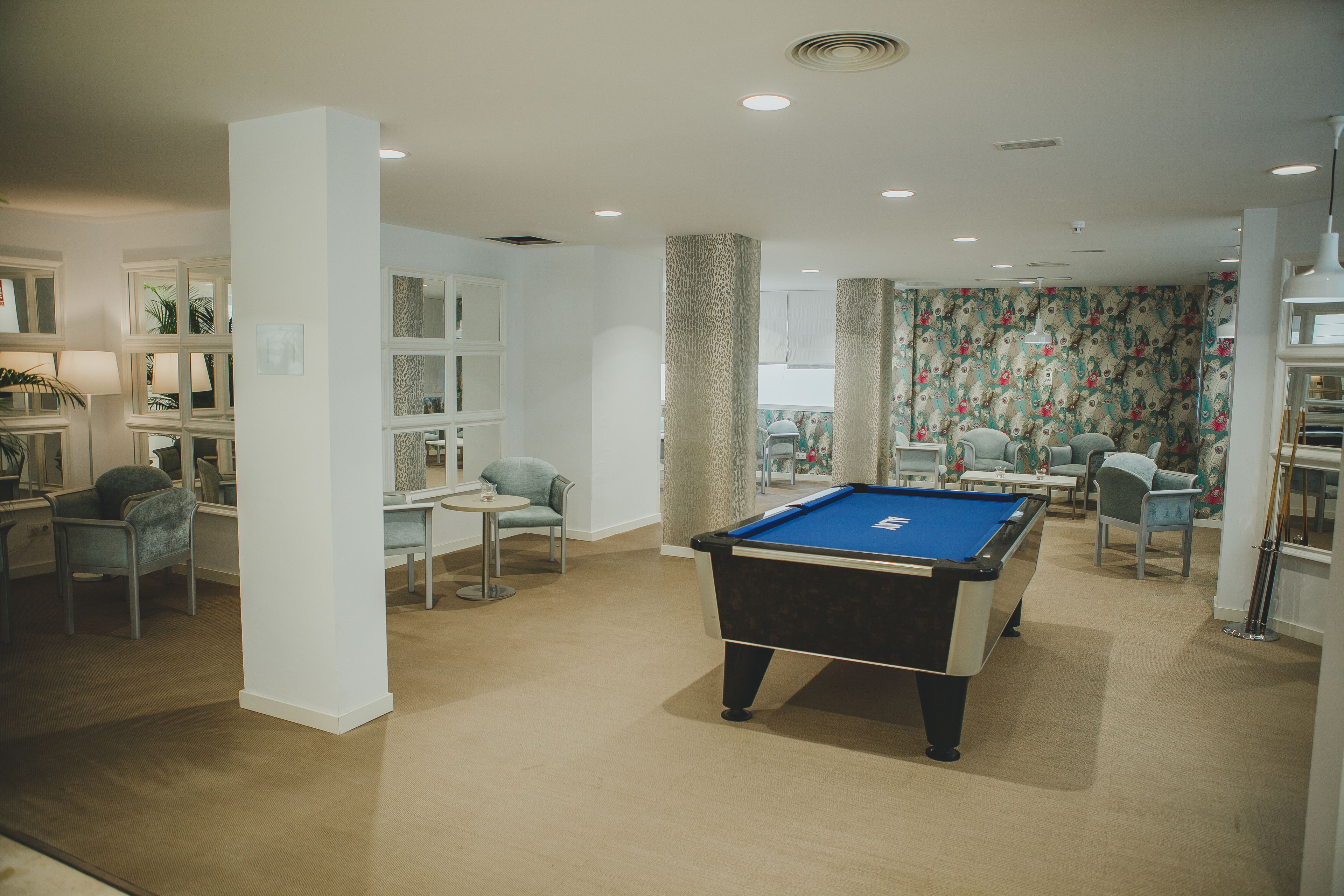 A pool table in a well-lit room with a carpeted floor, surrounded by chairs and a bookshelf.