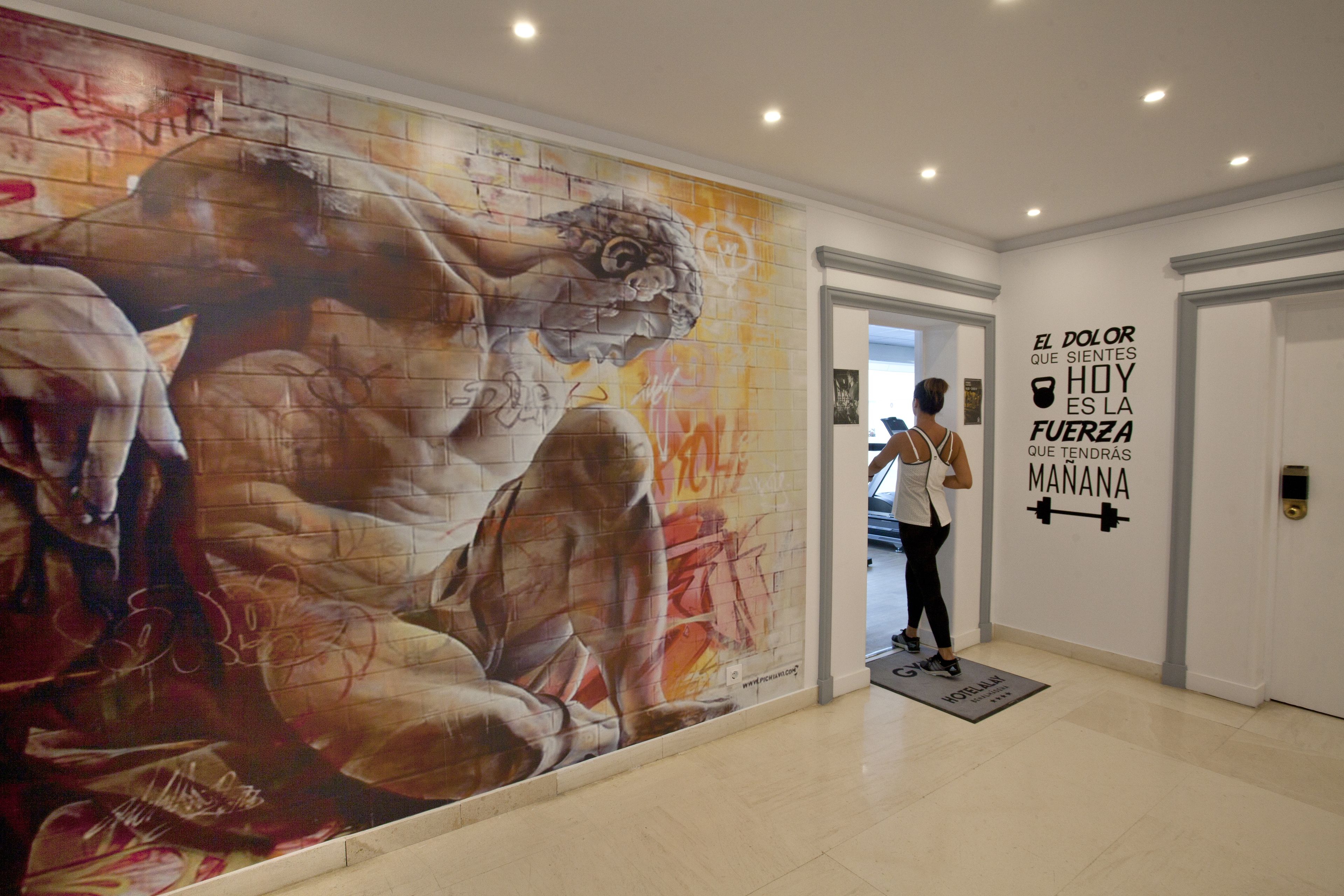 Un pasillo con un gran mural de una figura musculosa, una cita motivacional en la pared y una persona de pie cerca de la puerta.