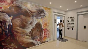 Un pasillo con un gran mural de una figura musculosa, una cita motivacional en la pared y una persona de pie cerca de la puerta.