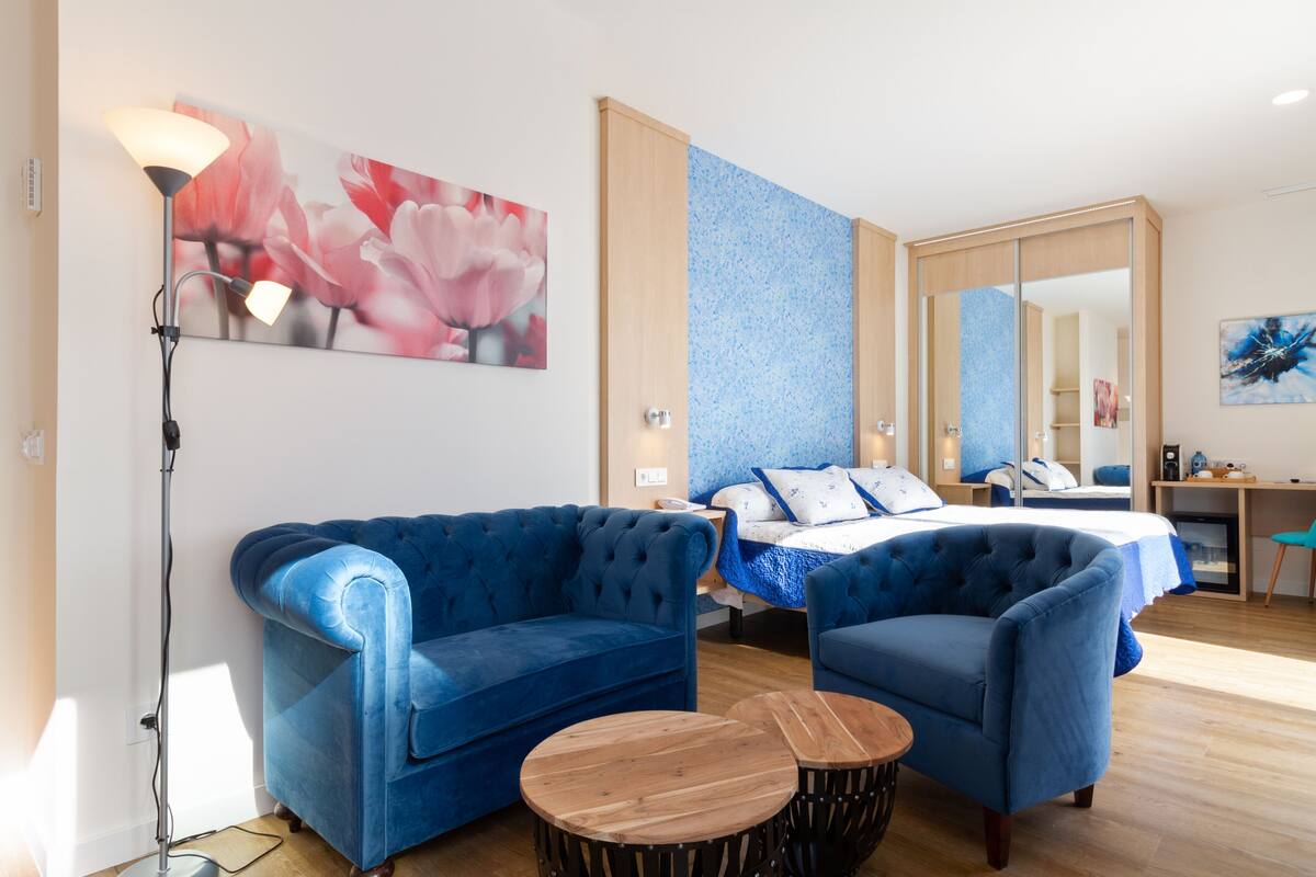 A modern hotel room with a blue sofa, a bed with a blue and white bedspread, a desk with a chair, and a large mirror.