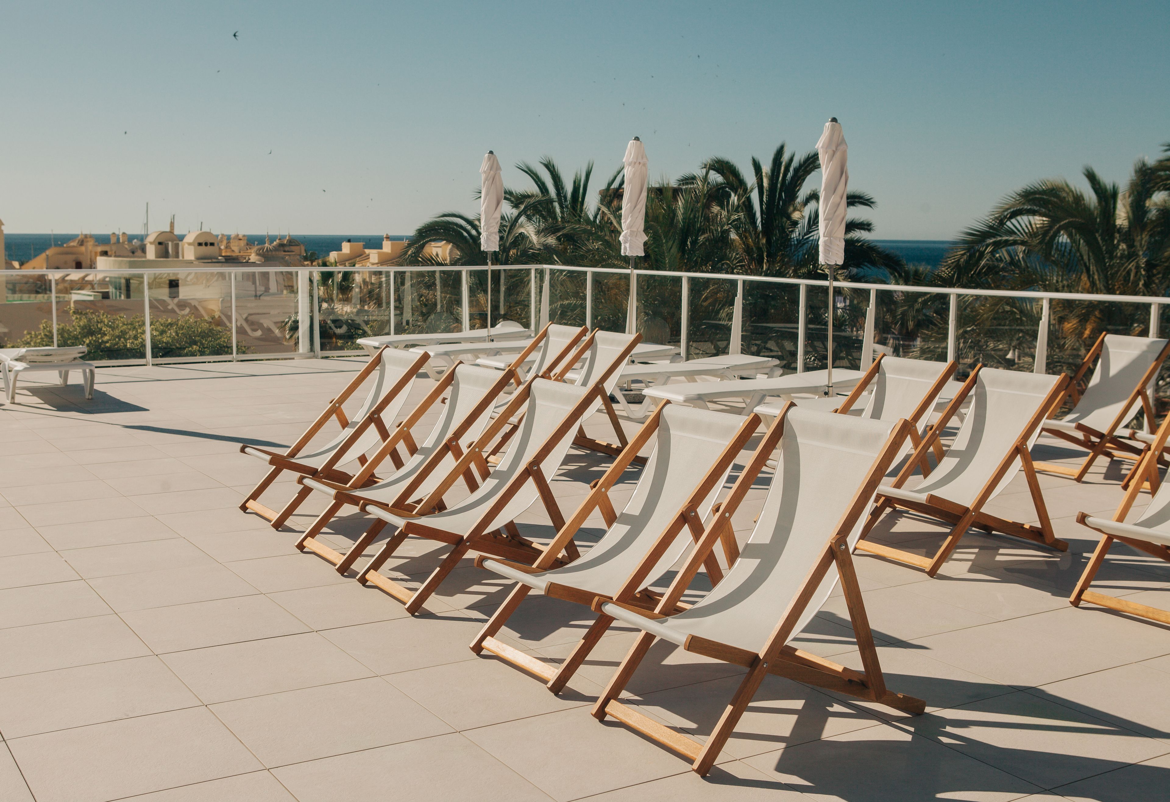 A rooftop terrace with sun loungers and umbrellas, overlooking a coastal area with palm trees and buildings.