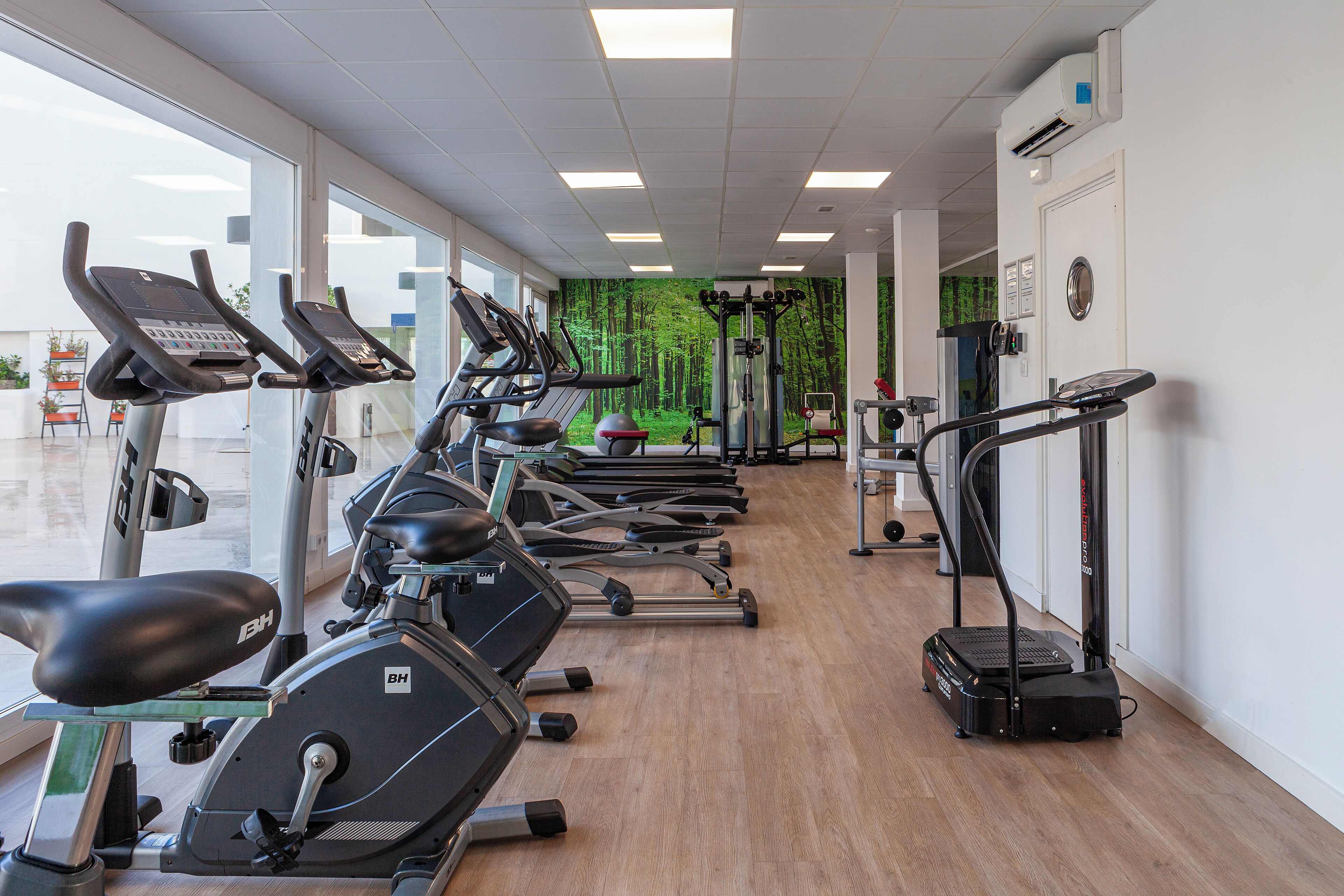 Un gimnasio con varias máquinas de ejercicio, piso de madera y ventanas amplias.