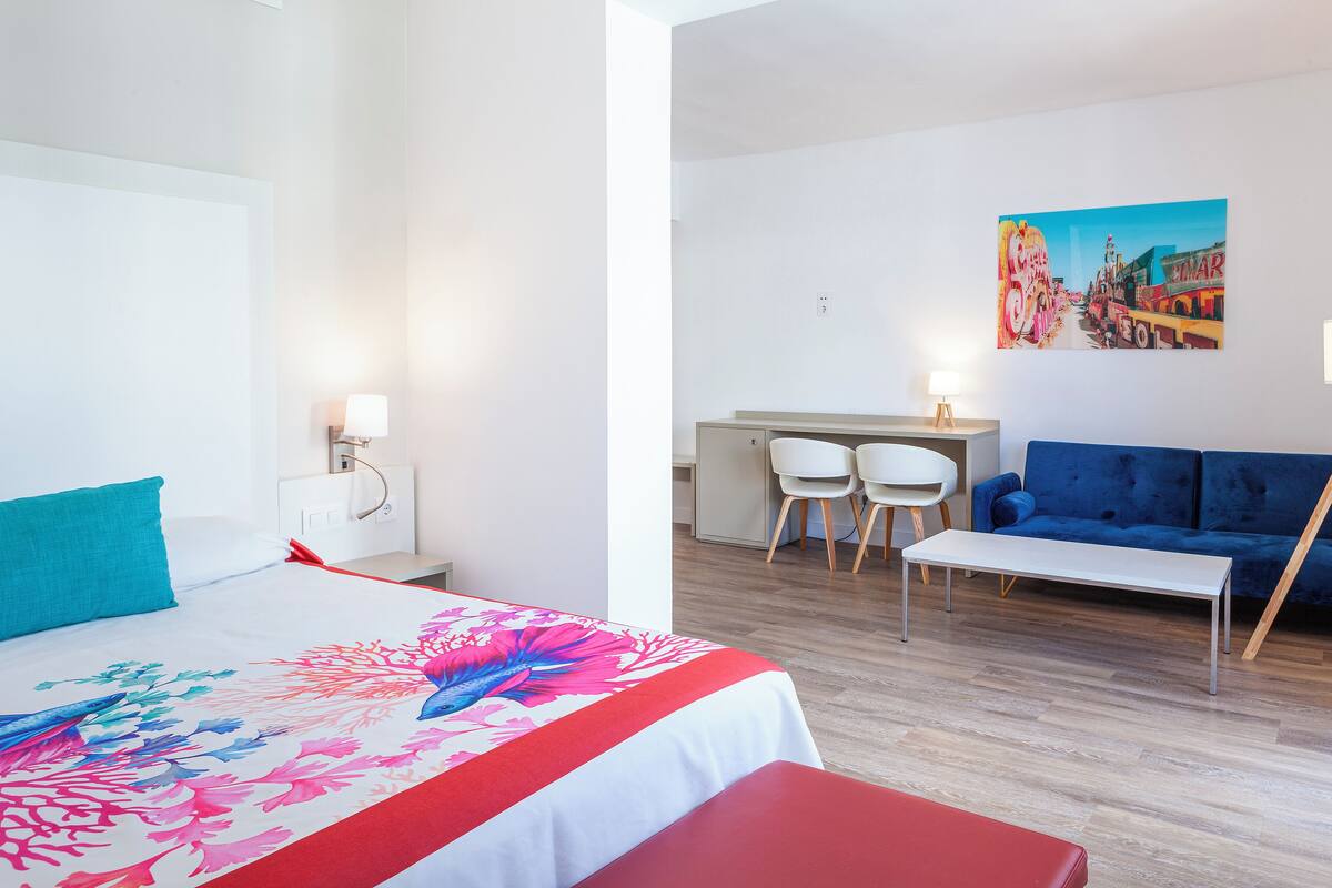 A hotel room with a bed, a desk, two chairs, a sofa, and a painting on the wall.