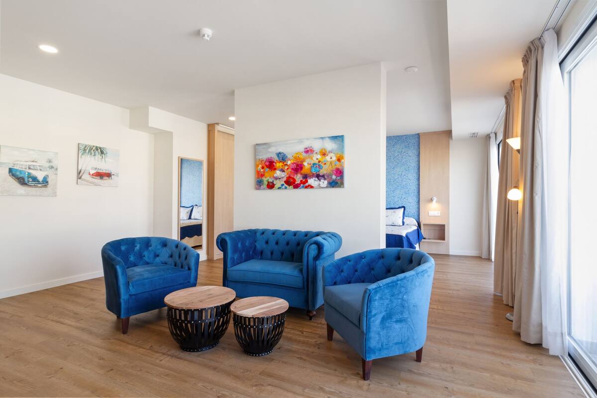 A modern hotel lobby with blue upholstered armchairs, a wooden coffee table, and a vibrant floral painting on the wall.