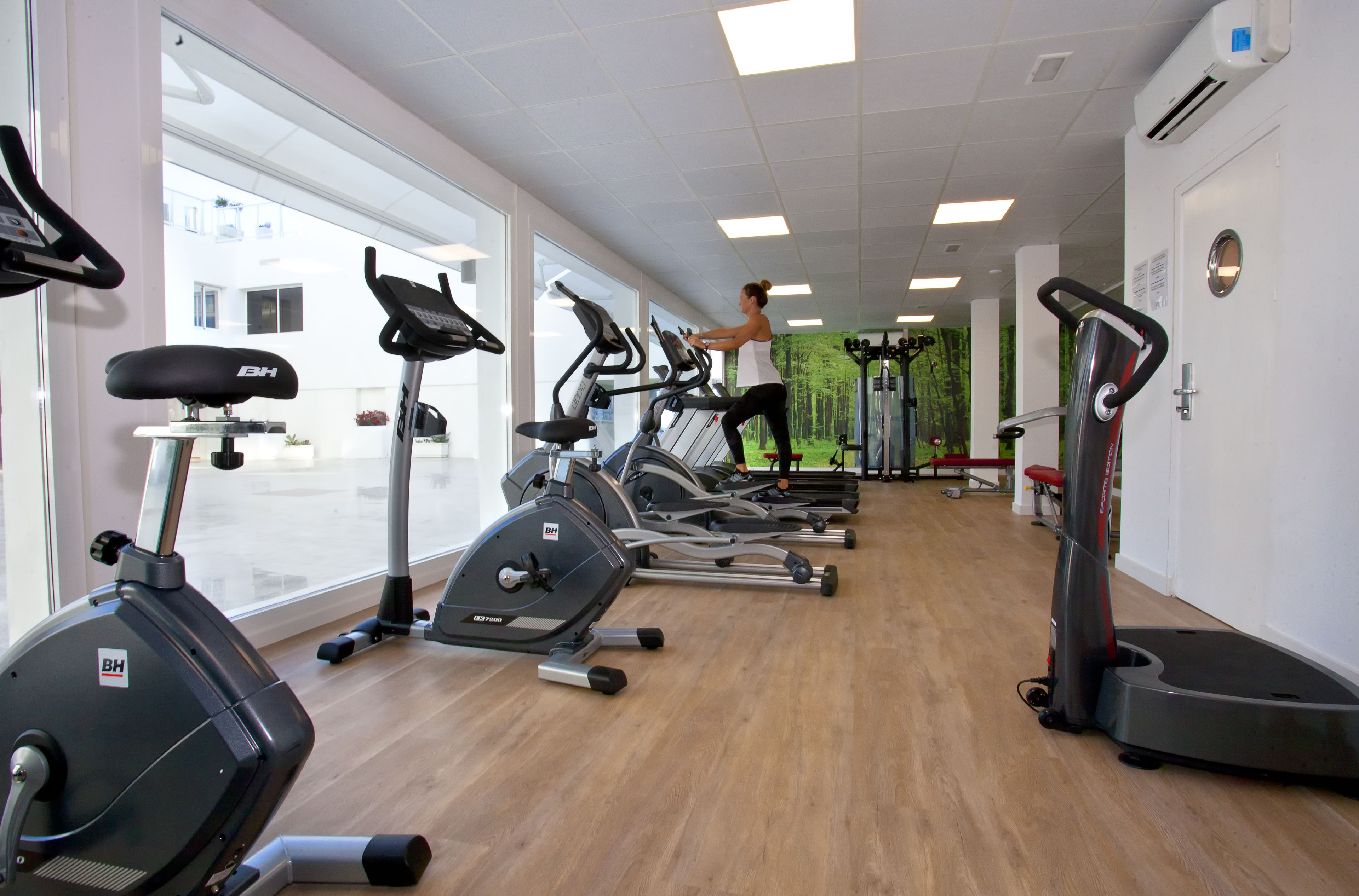 Un gimnasio con varias bicicletas estáticas y cintas de correr, ventanas amplias y suelo de madera.