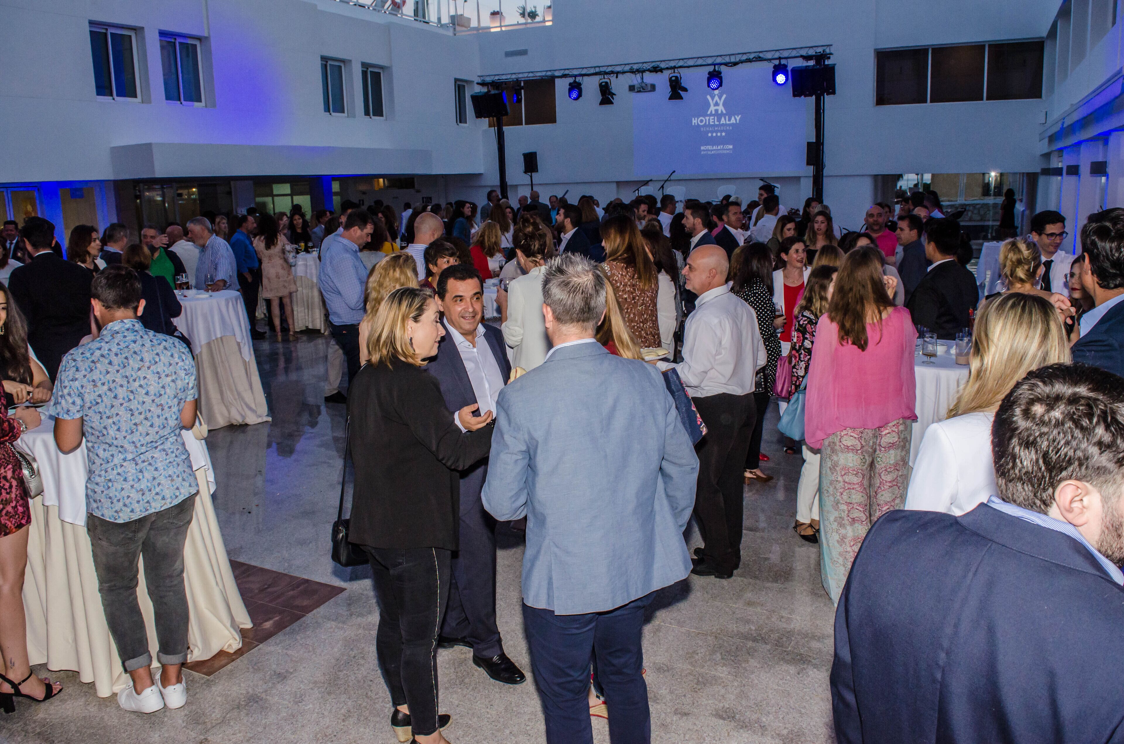 A crowded indoor event with people mingling at tables, a stage with a banner, and a modern building exterior.