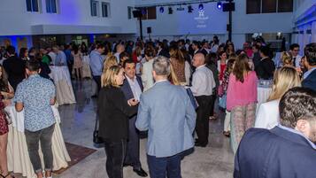 A crowded indoor event with people mingling at tables, a stage with a banner, and a modern building exterior.