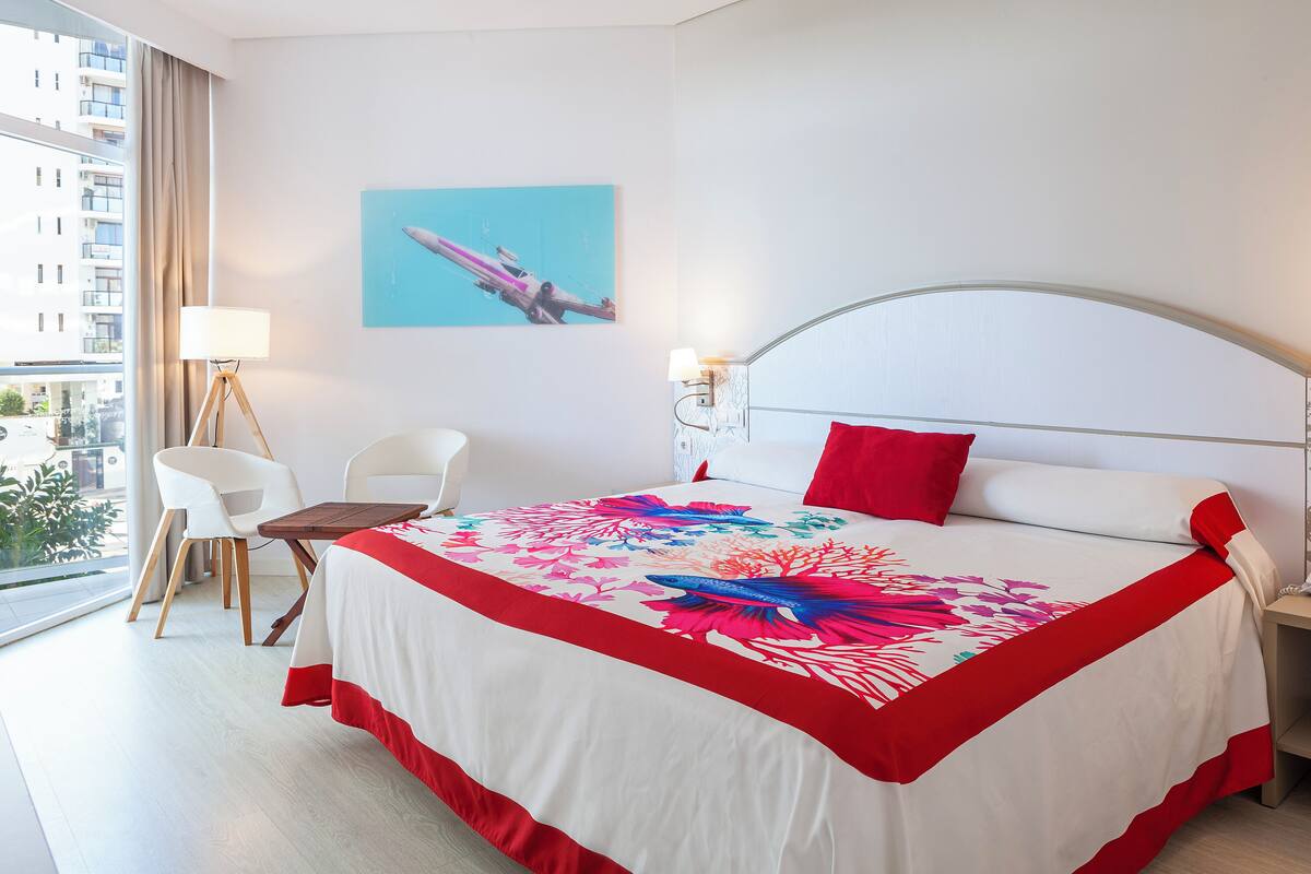 A hotel room with a large bed, a desk, two chairs, a lamp, and a city view.