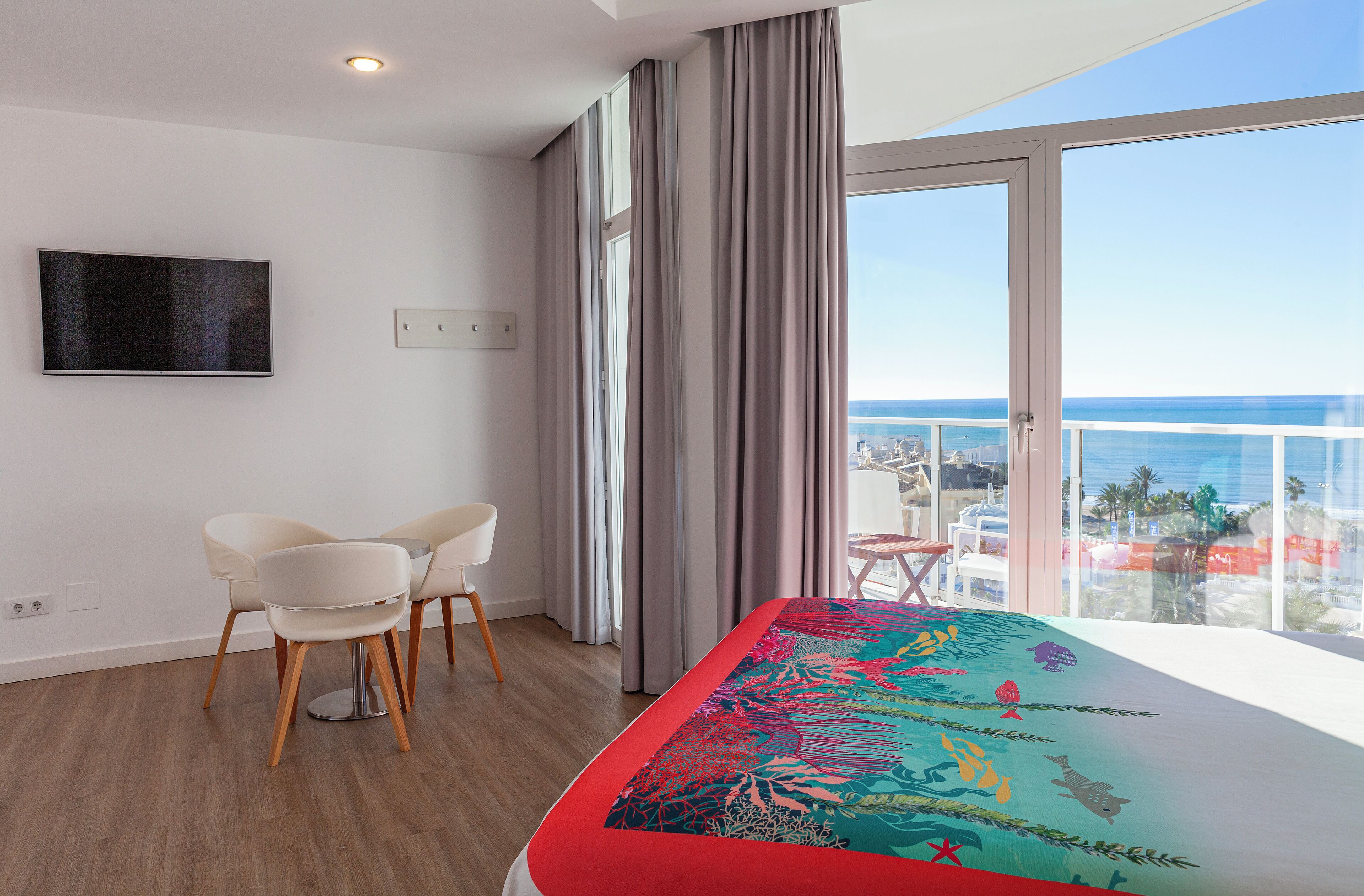Una habitación de hotel moderna con balcón, una cama con colcha colorida, televisión y zona de comedor con sillas.