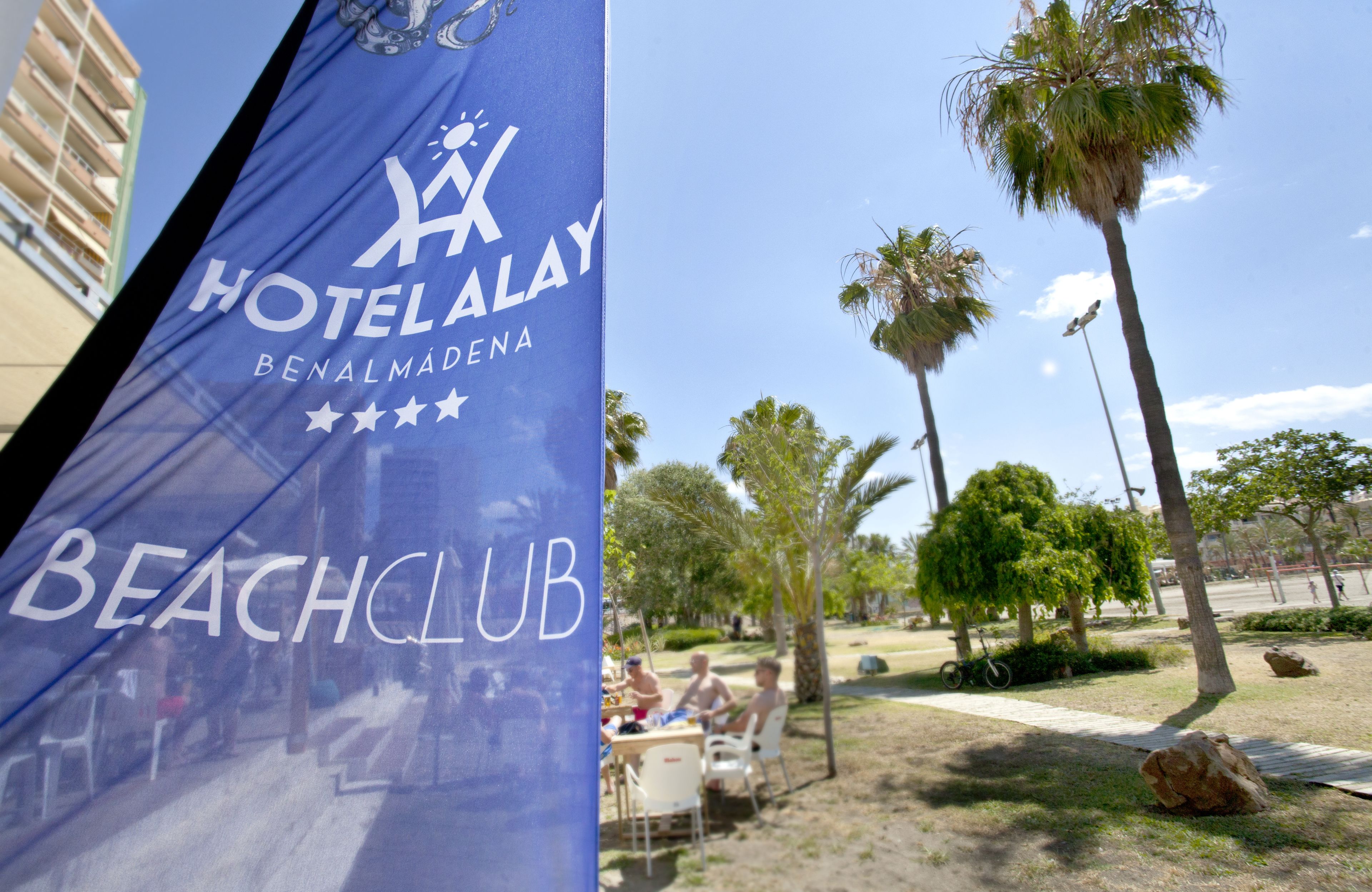 Una bandera azul con la inscripción "HOTEL ALAY BENALMADENA BEACH CLUB" y palmeras al fondo.
