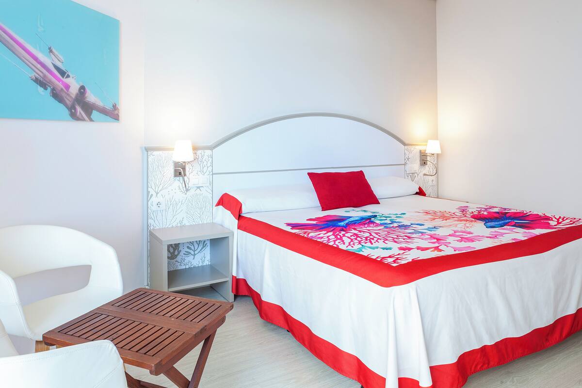 A hotel room with a bed, a nightstand, a chair, a table, and a wall with a painting.