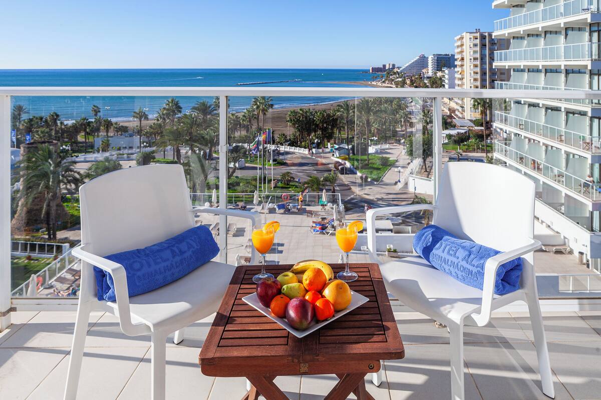 A balcony with two white chairs, a table with fruit and drinks, and a view of the beach and cityscape.
