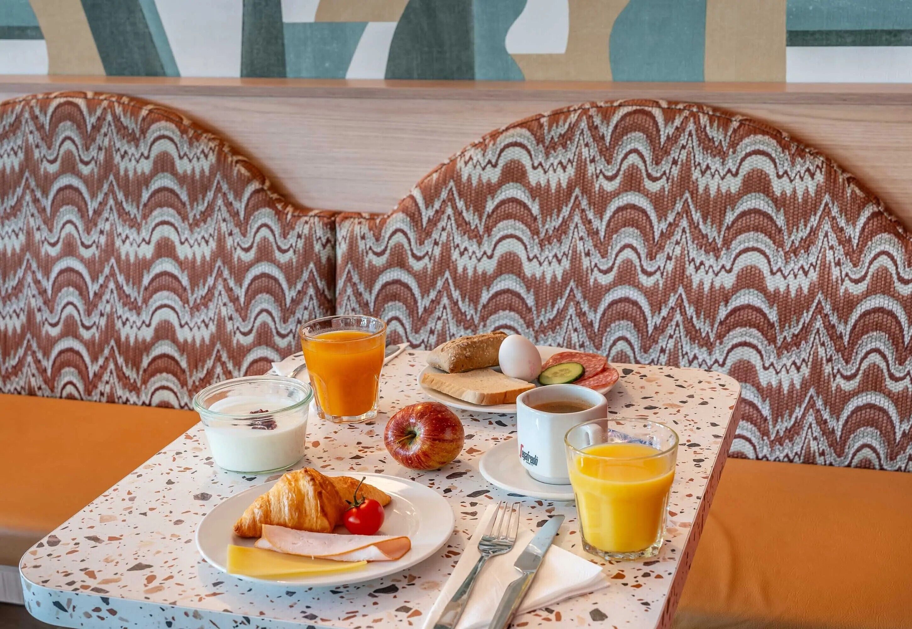 A breakfast setup on a table with croissants, ham, cheese, and a croissant with cherry tomatoes. There are two glasses of orange juice, a glass of milk, a cup of coffee, and a plate with eggs, tomatoes, and cucumber slices.