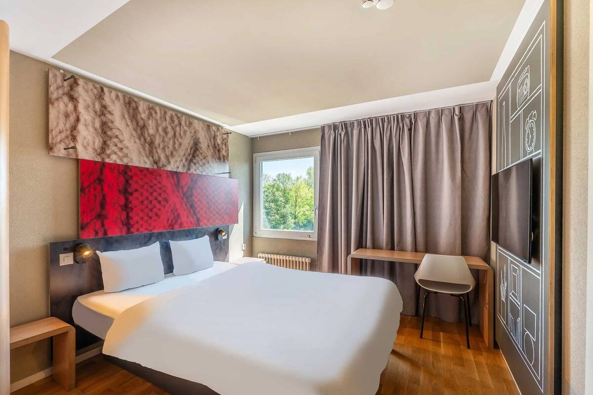 A hotel room with a large bed, a desk, a chair, a TV, and a window with curtains.