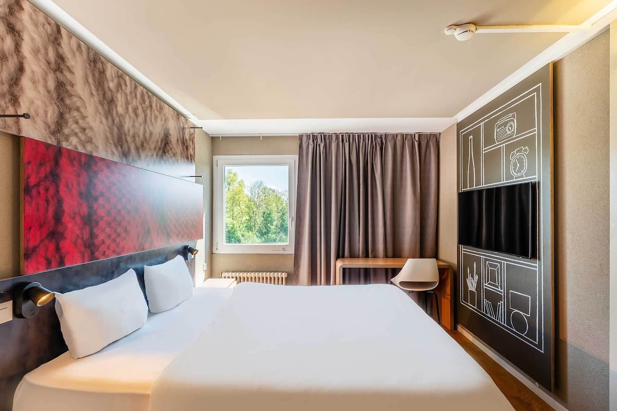 A modern hotel room with a large bed, a view of trees through the window, a wall-mounted TV, and a bedside table with a lamp.