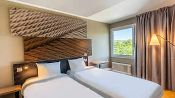 A hotel room with two beds, a large window with curtains, a radiator, and a wall-mounted lamp.