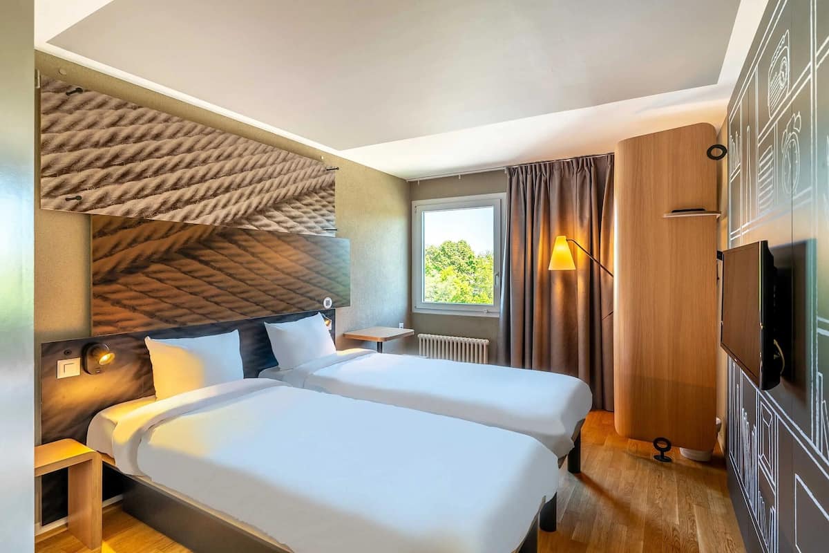 A hotel room with two beds, a small table, a TV, and a window with curtains.