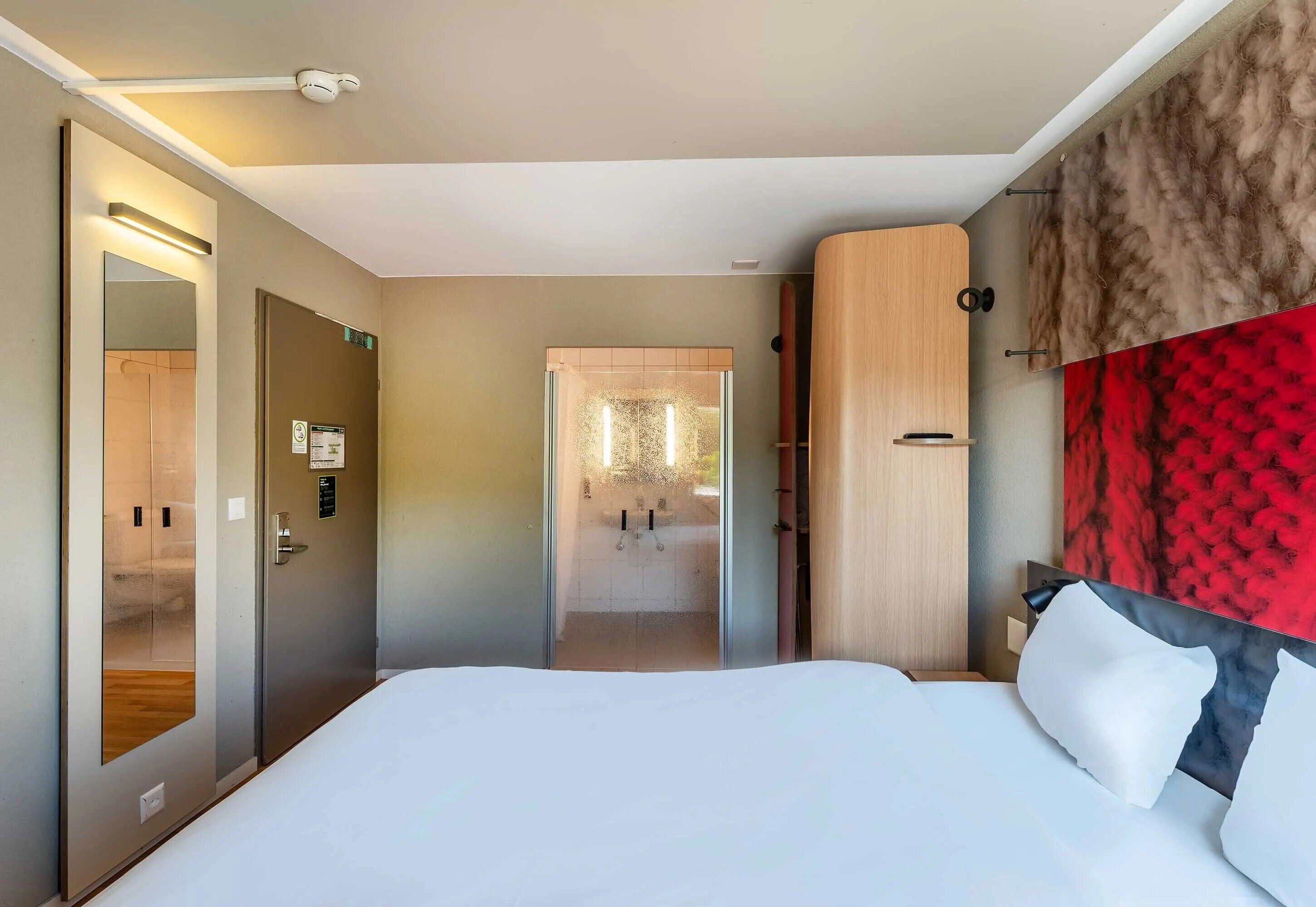 A modern hotel room with a large bed, a wooden headboard, a mirror, and a view of the bathroom.