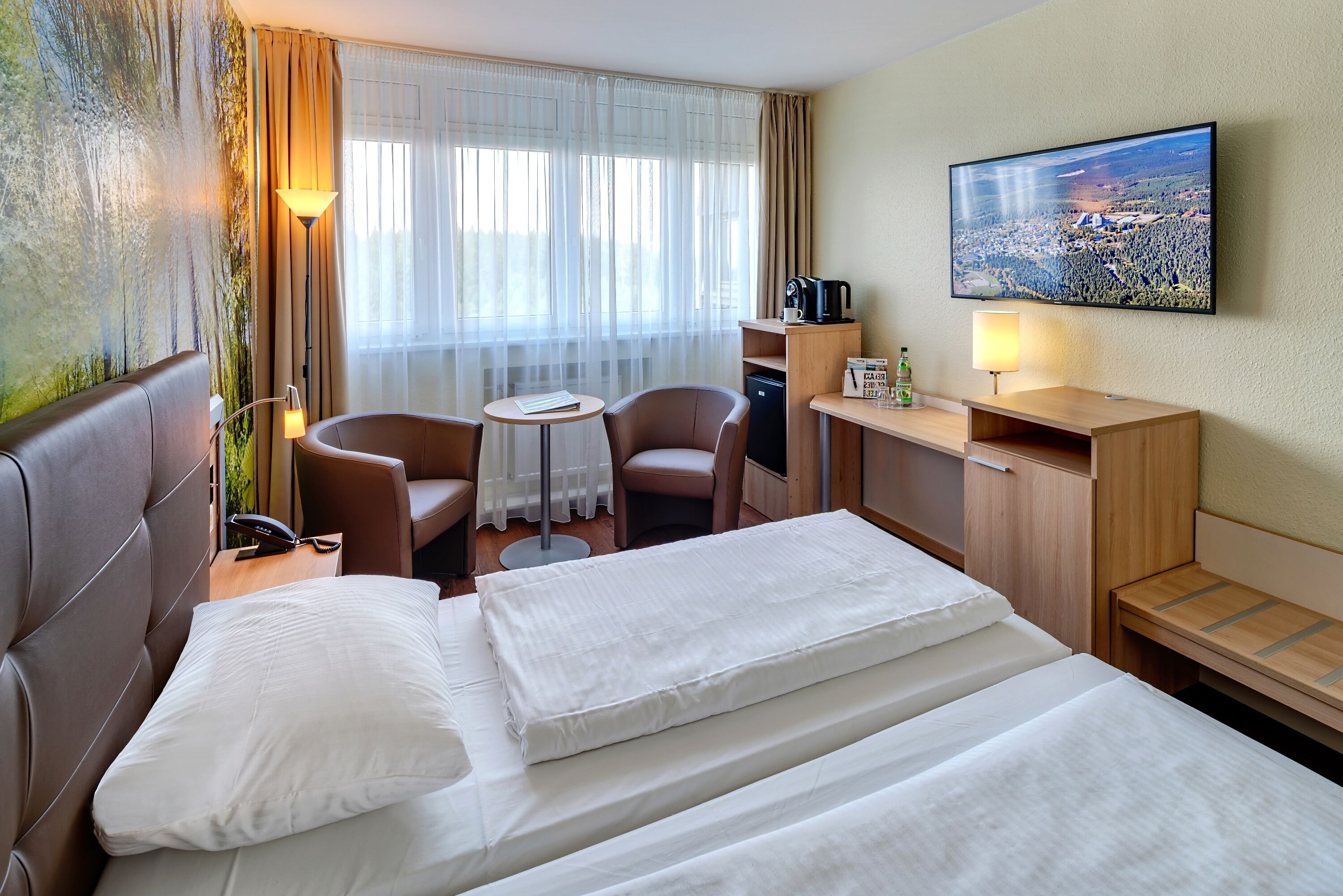 Ein Hotelzimmer mit einem Bett, zwei Stühlen, einem kleinen Tisch, einem Schreibtisch, einem Fernseher und einem Fenster mit Vorhängen.