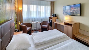 Ein Hotelzimmer mit einem Bett, zwei Stühlen, einem kleinen Tisch, einem Schreibtisch, einem Fernseher und einem Fenster mit Vorhängen.