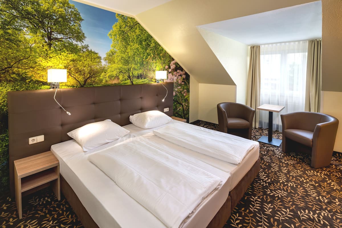 A hotel room with a large bed, a desk, two chairs, and a view of trees through a window.