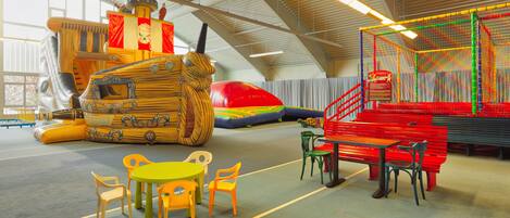 A spacious indoor play area with a large wooden ship play structure, colorful climbing net, and a red slide. There are several tables and chairs arranged for seating.