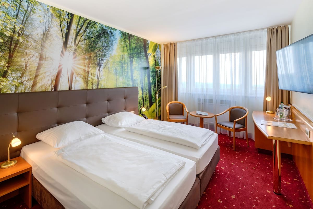 A hotel room with two beds, a desk, a chair, a TV, and a large forest mural on the wall.